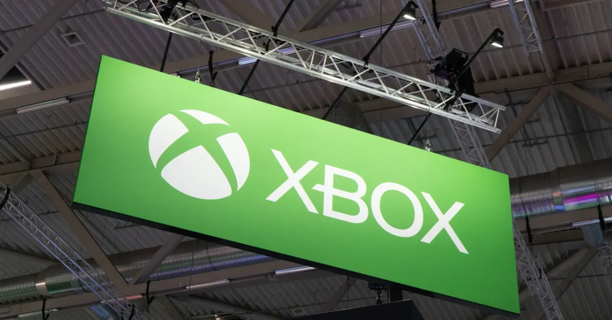 Grünes Xbox-Schild mit weißem Logo, aufgehängt in einer Messehalle mit sichtbarer Deckenkonstruktion und Beleuchtung.